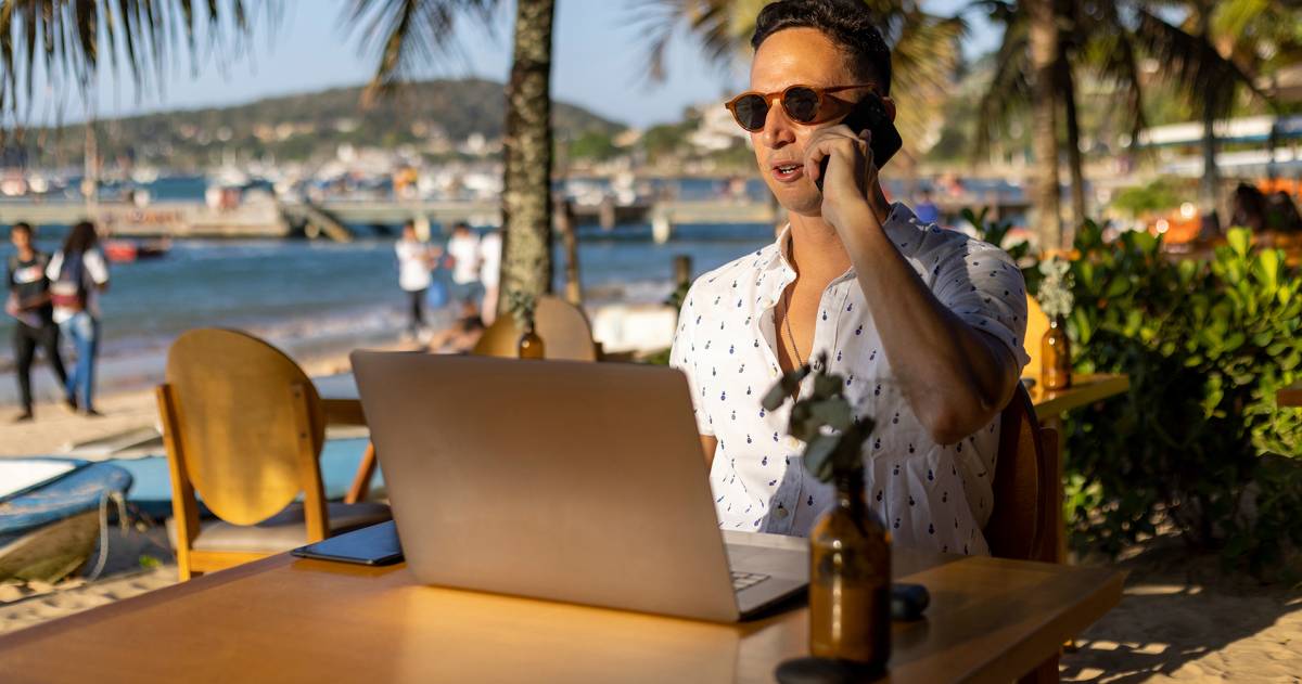 Zakenman zit op het strand achter zijn laptop en is aan het bellen